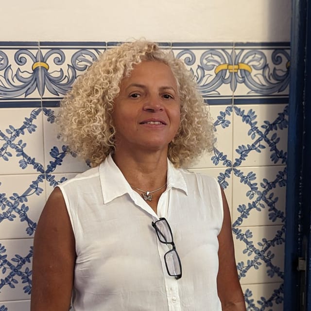 Jô em seu ateliê de costura, trabalhando com precisão em ajustes de roupas com ferramentas profissionais e tecidos ao fundo